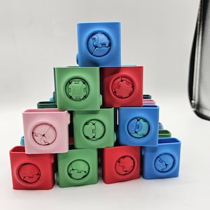 May include: A pyramid of colorful, square-shaped containers in red, green, blue, and pink. Each container features a unique, circular design on its front. The containers are stacked on a white surface.