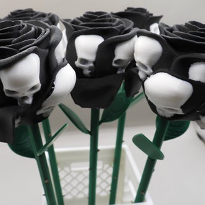 May include: A bouquet of six black roses with white skull designs. The roses are arranged in a white vase with a green grid pattern.