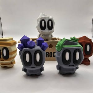 May include: A group of colourful, stylised figurines with large oval eyes. The figures are in various colours, including grey, orange, and tan, and some have decorative elements like crystals or a leafy headpiece. The word "ROC" is visible on a box.