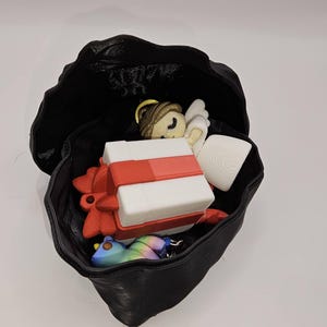 May include: A black, open, leather-like container filled with various items. Inside are a white gift box with a red ribbon, a small angel figurine, a white scroll, and a colorful bird-shaped keychain.
