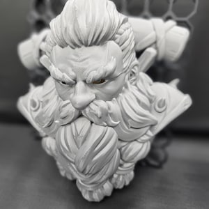 May include: A detailed, gray bust of a bearded warrior with a stern expression. The sculpture features intricate details in the hair, beard, and armor, with visible texture. The eyes are a contrasting brown color.