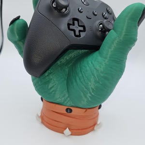 May include: A green, 3D-printed hand-shaped gaming controller holder. The hand is holding a black controller. The base of the holder is orange with white teeth-like accents. The controller has a cross-shaped directional pad.
