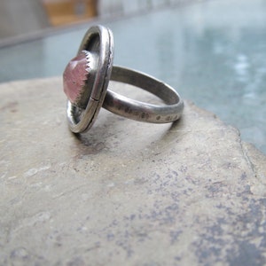 May include: A silver ring with a pink gemstone set in a bezel setting. The ring is made of a thick band of metal and has a textured finish.