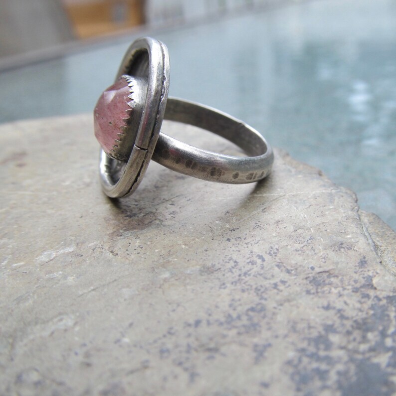 May include: A silver ring with a pink gemstone set in a bezel setting. The ring is made of a thick band of metal and has a textured finish.