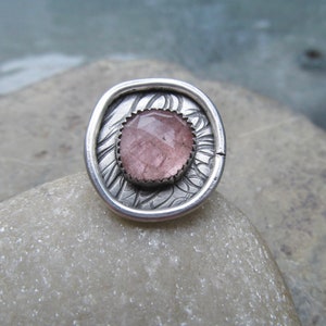 May include: A silver ring with a pink gemstone set in a textured, ornate design.