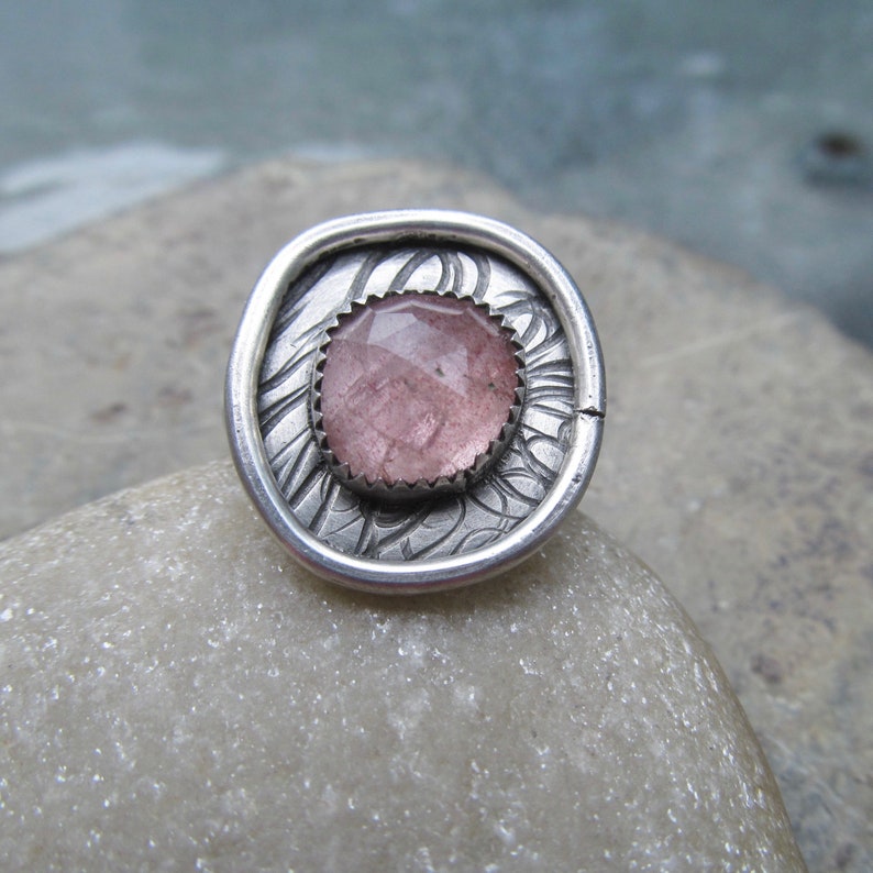 May include: A silver ring with a pink gemstone set in a textured, ornate design.