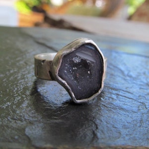 May include: A silver ring with a dark gray geode stone set in the center. The stone has a rough, natural shape and is surrounded by a silver band.