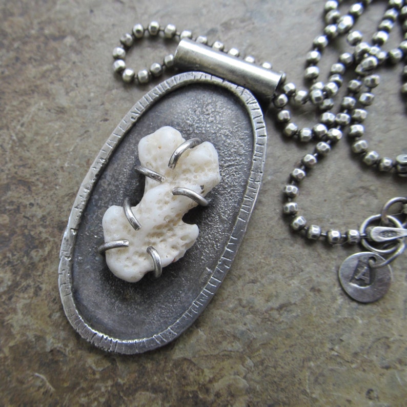May include: A silver pendant necklace with a white coral piece encased in a textured oval frame. The chain is a silver ball chain.