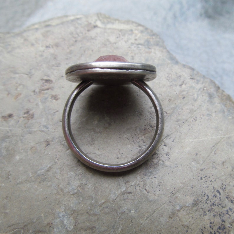 May include: A silver ring with a pink gemstone set in a bezel setting. The ring is on a gray background.