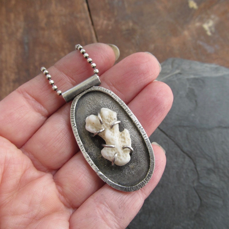 May include: A silver pendant necklace with a white coral piece encased in a silver oval frame. The pendant has a textured edge and a silver chain.