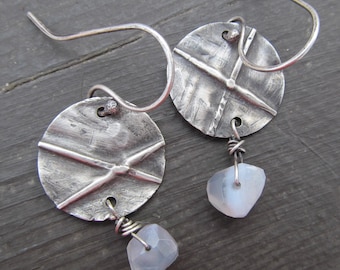 May include: Silver earrings with a textured circular design and a dangling moonstone. The earrings have a hammered metal look and are a unique and stylish accessory.