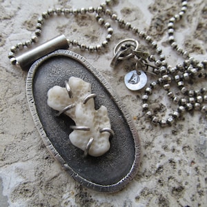 May include: A silver necklace with a pendant featuring a white coral piece encased in a silver oval frame. The necklace has a silver chain with a small silver charm with the letter 'A' on it.