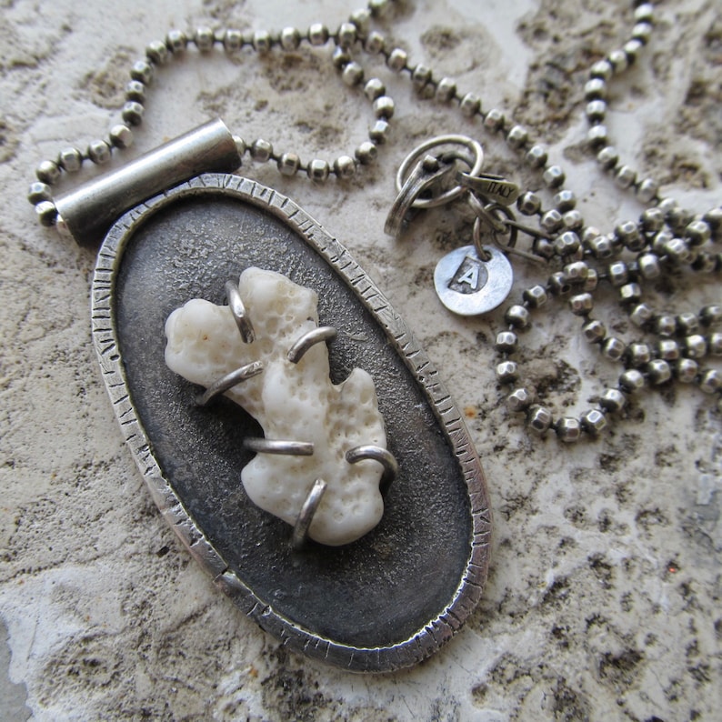 May include: A silver necklace with a pendant featuring a white coral piece encased in a silver oval frame. The necklace has a silver chain with a small silver charm with the letter 'A' on it.