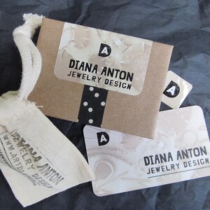 May include: A brown paper bag with a white sticker that says "DIANA ANTON JEWELRY DESIGN" and a black and white polka dot pattern. There is a smaller white sticker with the letter "A" on the bag. There is a white card with the same text and a black and white image of gears.