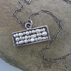 May include: A silver chain necklace with a rectangular pendant featuring a row of white pearls set in silver wire.