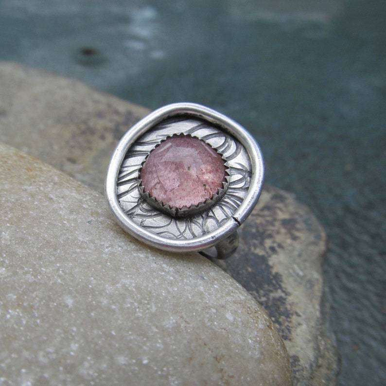 May include: A silver ring with a pink and white gemstone set in a circular bezel. The ring has a textured design around the stone.
