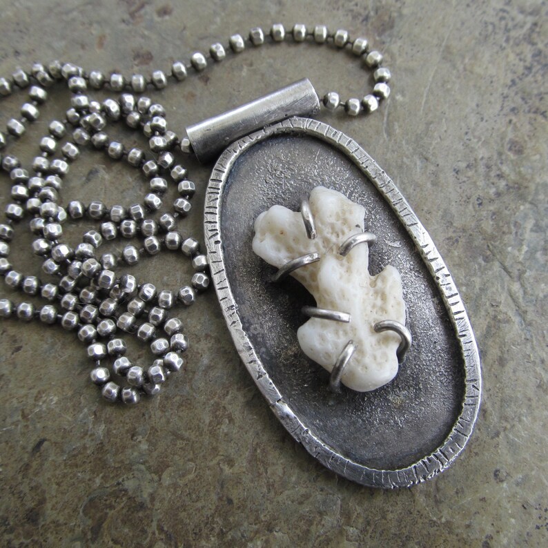 May include: A silver necklace with a pendant featuring a white coral piece encased in a textured oval frame.