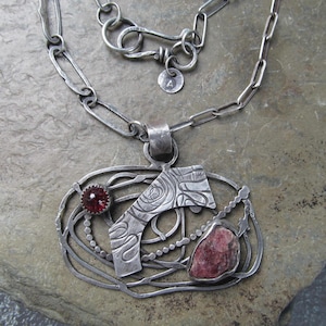 May include: A handcrafted silver necklace featuring a large pendant with a textured design and a red gemstone. The pendant is connected to a chain with a unique clasp. The necklace is displayed on a stone surface.