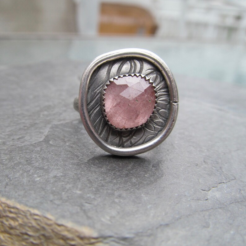 May include: A silver ring with a pink gemstone set in a textured, ornate design.