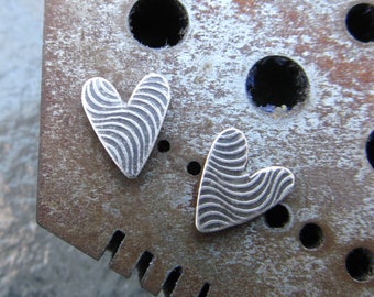 May include: Two silver heart-shaped earrings with a wavy, textured design. The earrings are displayed on a metal surface.
