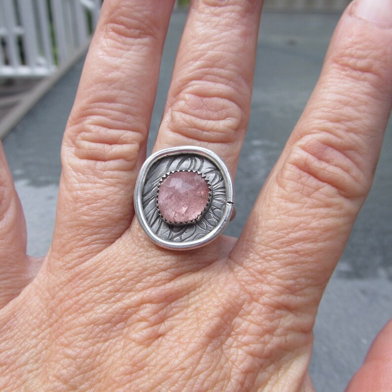 May include: A silver ring with a pink gemstone set in a textured, floral design.