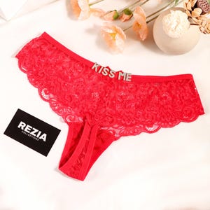 May include: Bright red lace panties featuring a scalloped edge and a silver-toned rhinestone embellishment on the front waistband that spells out the text KISS ME. A small black rectangular card with the white text REZIA COLLECTIONS is included.
