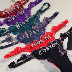Pode incluir: Uma coleção de tangas em várias cores, incluindo vermelho, roxo, azul-petróleo, azul e preto. Cada peça apresenta acabamento em renda festonada, alças ajustáveis com detalhes dourados e texto com incrustações de strass: BITCH, KISS ME, MISS, BABY, FUCK ME e CUSTOM.