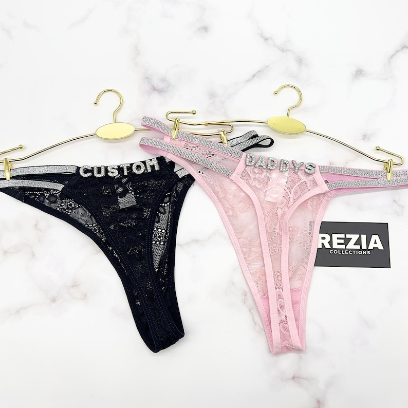 May include: Two thong panties made of floral lace, one black and one light pink. Each features double silver glitter straps on the sides and a silver rhinestone word across the front waistband. The black pair reads CUSTOM and the pink pair reads DADDYS.