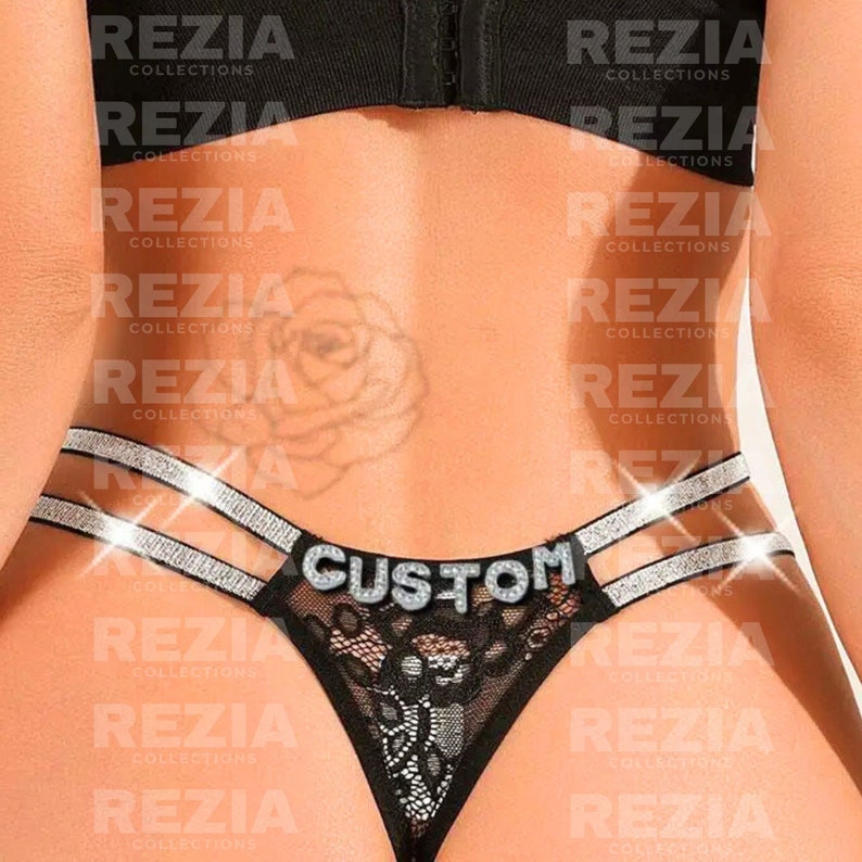 May include: Black lace thong featuring double silver glittery straps on each side and the word CUSTOM spelled out in silver rhinestone letters across the center. The garment has a black elastic waistband and a sheer black lace front panel.