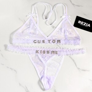 May include: A white lingerie set with floral lace detailing. The bra and the waist band of the bottoms have the text "CUSTOM" and "KISS ME" spelled out in rhinestones. The set includes a triangle bra, a thong, and a matching waist band.