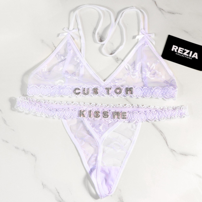 May include: A white lingerie set with floral lace detailing. The bra and the waist band of the bottoms have the text "CUSTOM" and "KISS ME" spelled out in rhinestones. The set includes a triangle bra, a thong, and a matching waist band.