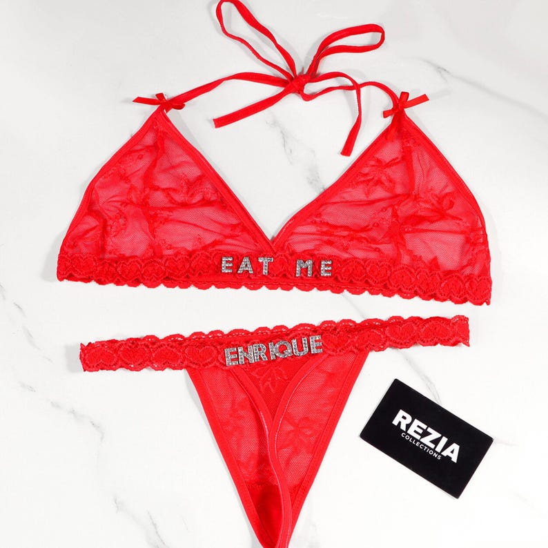May include: Red lingerie set featuring a triangle bralette and a thong. The bralette has the text "EAT ME" and the thong has the text "ENRIQUE" in rhinestones. The set includes a small black card with the text "REZIA COLLECTIONS".