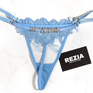 May include: Pale blue thong underwear with a detailed lace pattern and the words "I'M YOURS" in silver lettering. The underwear has adjustable straps and a sheer section. A black card with "REZIA COLLECTIONS" is visible.