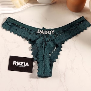 May include: Dark green lace thong featuring a scalloped edge and a central rhinestone embellishment spelling the word DADDY. A small black rectangular card with the white text REZIA COLLECTIONS is positioned next to the garment.