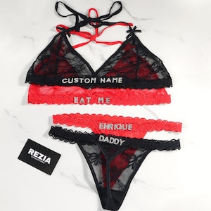 May include: Black and red lingerie set with lace detailing. The set includes a bralette and a thong, with the words "CUSTOM NAME", "EAT ME", "ENRIQUE" and "DADDY" in rhinestones. A "REZIA COLLECTIONS" card is also visible.