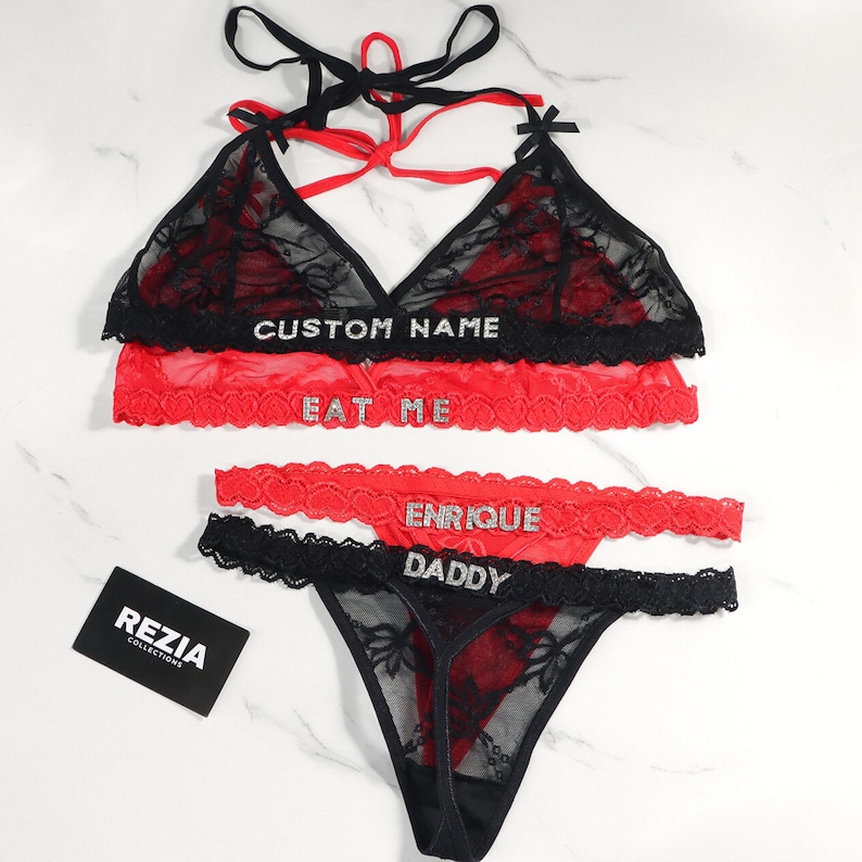 May include: Black and red lingerie set with lace detailing. The set includes a bralette and a thong, with the words "CUSTOM NAME", "EAT ME", "ENRIQUE" and "DADDY" in rhinestones. A "REZIA COLLECTIONS" card is also visible.