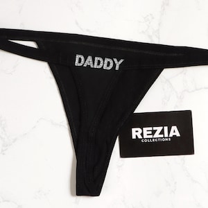 Pode incluir: Tanga preto com a palavra "DADDY" em glitter prateado. A roupa íntima está sobre uma superfície de mármore branco. Um cartão de visita preto com "REZIA COLLECTIONS" está ao lado.
