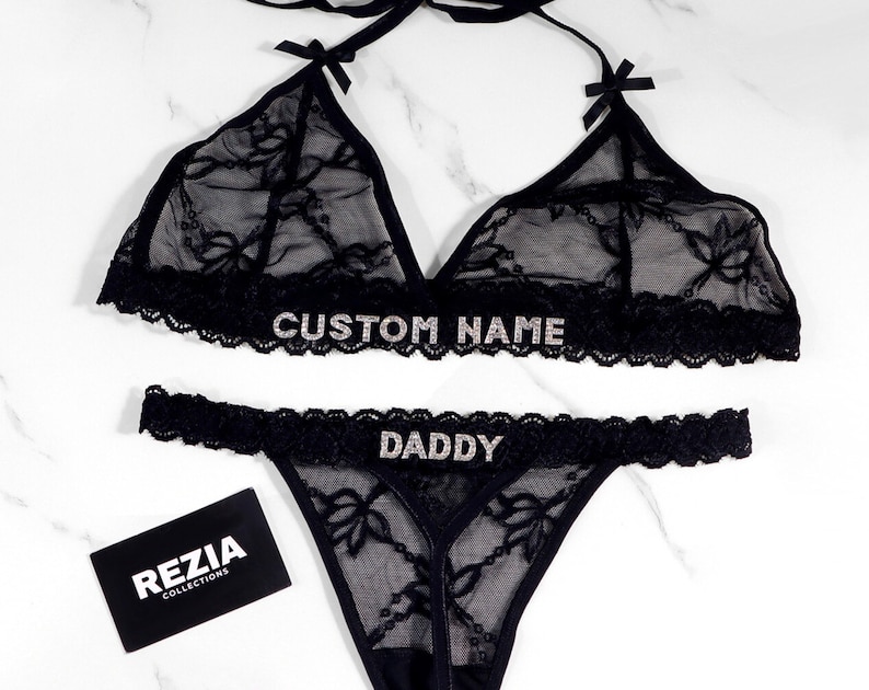 May include: Black lace lingerie set with the text "CUSTOM NAME" on the top and "DADDY" on the bottom. The set includes a halter top with bow accents and a thong. A black business card with the text "REZIA COLLECTIONS" is also in the image.