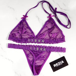 May include: A purple lingerie set featuring a halter-style bra and a thong. The bra and thong are adorned with the word "LIBRA" in silver rhinestones. The set is made of sheer fabric with floral embroidery and lace trim. A black and white business card is in the image.