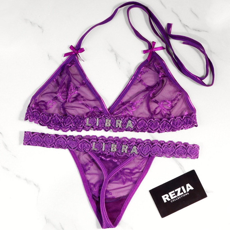 May include: A purple lingerie set featuring a halter-style bra and a thong. The bra and thong are adorned with the word "LIBRA" in silver rhinestones. The set is made of sheer fabric with floral embroidery and lace trim. A black and white business card is in the image.