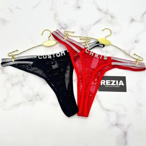 May include: Two thongs hanging on gold hangers, one black and one red, both featuring lace fabric and silver glittery side straps. The black thong has the word CUSTOM in rhinestone letters, and the red thong has the word CHRIS in rhinestone letters.