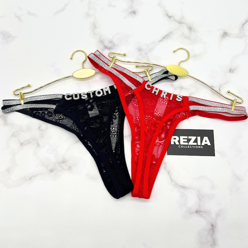 May include: Two thongs hanging on gold hangers, one black and one red, both featuring lace fabric and silver glittery side straps. The black thong has the word CUSTOM in rhinestone letters, and the red thong has the word CHRIS in rhinestone letters.