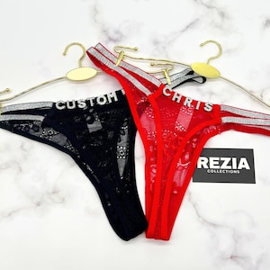 Könnte beinhalten: Zwei Tangas, einer in Schwarz und einer in Rot, aus Spitzenstoff mit silberfarbenen Glitzer-Seitenbändern. Jedes Paar ist mit Strassbuchstaben verziert, wobei der schwarze Tanga CUSTOM und der rote CHRIS zeigt, hängend an goldenen Kleiderbügeln.