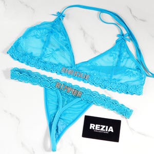May include: A turquoise lace lingerie set featuring a halter-style bra and a thong. The bra and thong are adorned with the name "ENRIQUE" and "RUMIER" in sparkling embellishments. A black card with "REZIA COLLECTIONS" is also present.