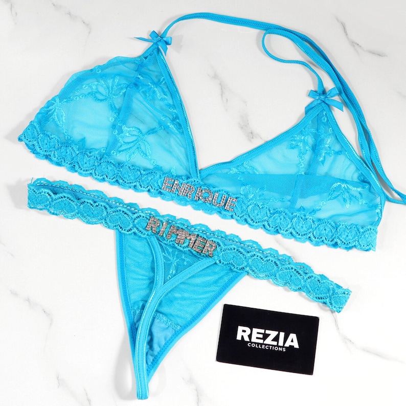 May include: A turquoise lace lingerie set featuring a halter-style bra and a thong. The bra and thong are adorned with the name "ENRIQUE" and "RUMIER" in sparkling embellishments. A black card with "REZIA COLLECTIONS" is also present.