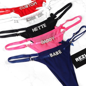 May include: A collection of thong underwear in red, white, black, pink, and navy blue. Each thong features a rhinestone embellishment with text: "CUSTOM", "KISS ME", "NETTE", "DADDY", and "BABE". The underwear has adjustable straps.