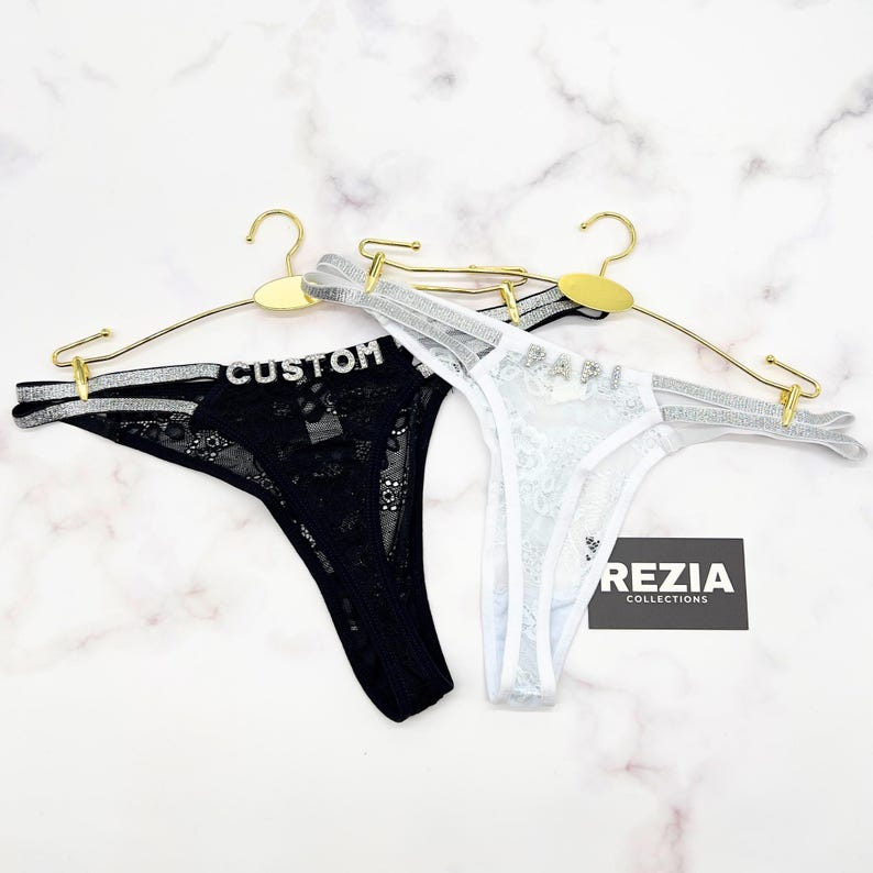 May include: A black lace thong with the word CUSTOM in rhinestones and a white lace thong with the word PAPI in rhinestones, both featuring silver glitter straps on gold hangers. A black tag displays the text REZIA COLLECTIONS in white.