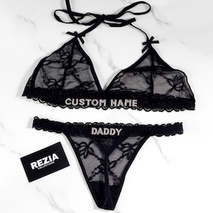 May include: Black lingerie set with a halter top and thong. The top and bottom are made of sheer lace with floral patterns and embellished with the text "CUSTOM NAME" and "DADDY". A small black card with the text "REZIA COLLECTIONS" is also present.