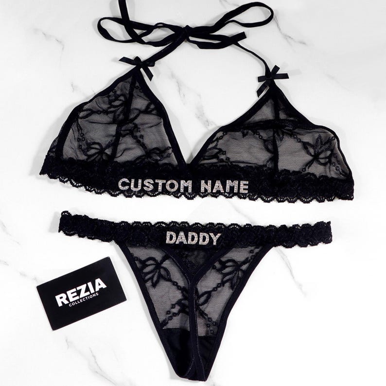 May include: Black lingerie set with a halter top and thong. The top and bottom are made of sheer lace with floral patterns and embellished with the text "CUSTOM NAME" and "DADDY". A small black card with the text "REZIA COLLECTIONS" is also present.