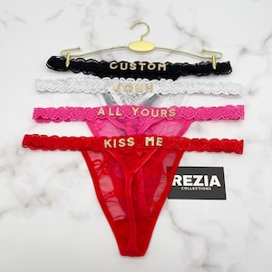 Może przedstawiać: Cztery stringi z czarnej, białej, r&oacute;żowej i czerwonej koronki, każdy z napisem ze złotych cyrkonii na pasku: CUSTOM, JOHN, ALL YOURS i KISS ME. Produkty są zawieszone na złotym metalowym wieszaku z metką REZIA COLLECTIONS.